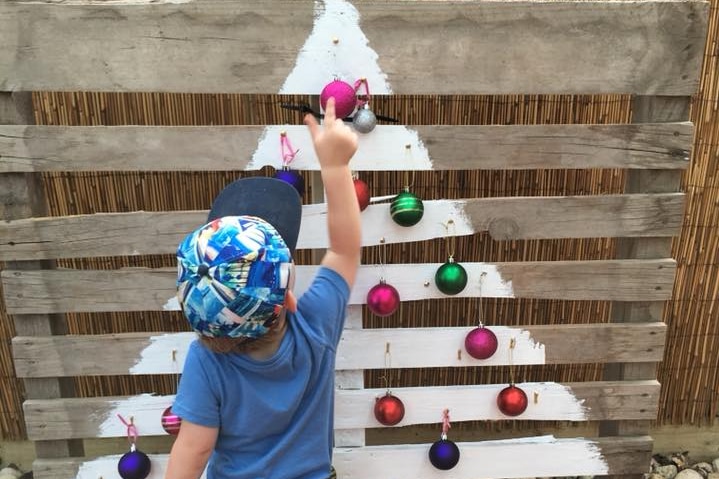A Christmas tree painted in white on a wooden pallet, hung with baubles. Child points to top bauble.