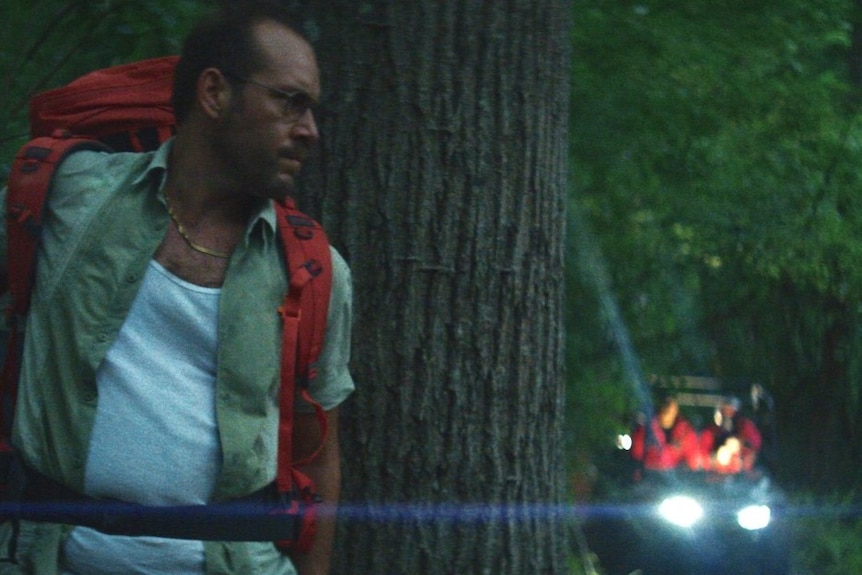 Dean Imperial, a middle aged man with a backpack his back against a tree in a dark night, in the film Lapsis