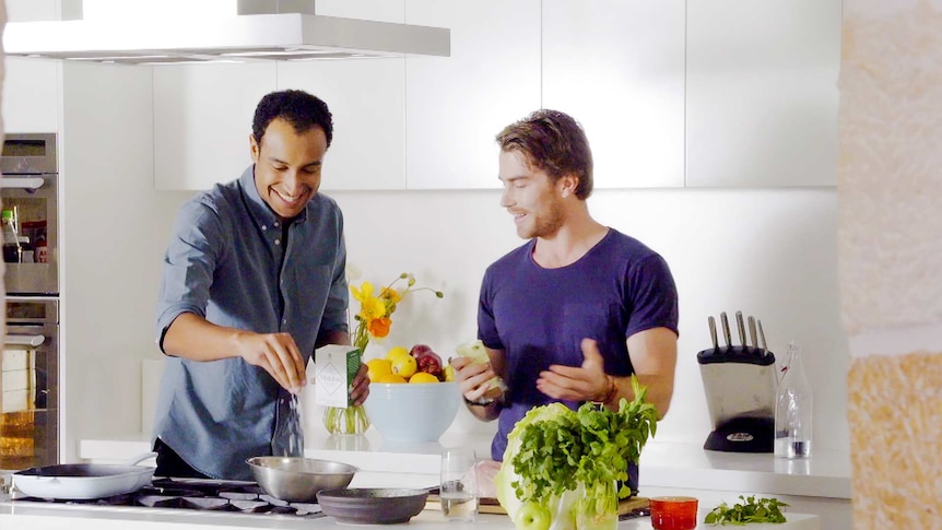Tall man with dark features sprinkles salt into a bowl which a man with blond hair gives him cooking tips alongside him.