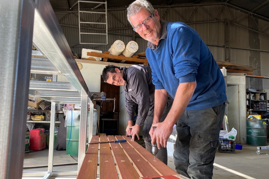 Two men are leaving over wooden planks with drills, smiling.