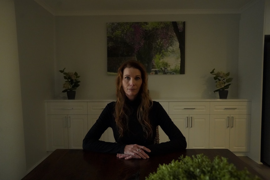 A woman in black sits at a table looking serious.