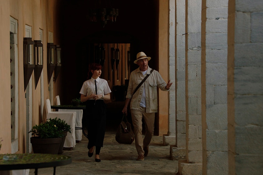 A woman in hospitality attire walks down corridor with gesticulating man with Panama sun hat.
