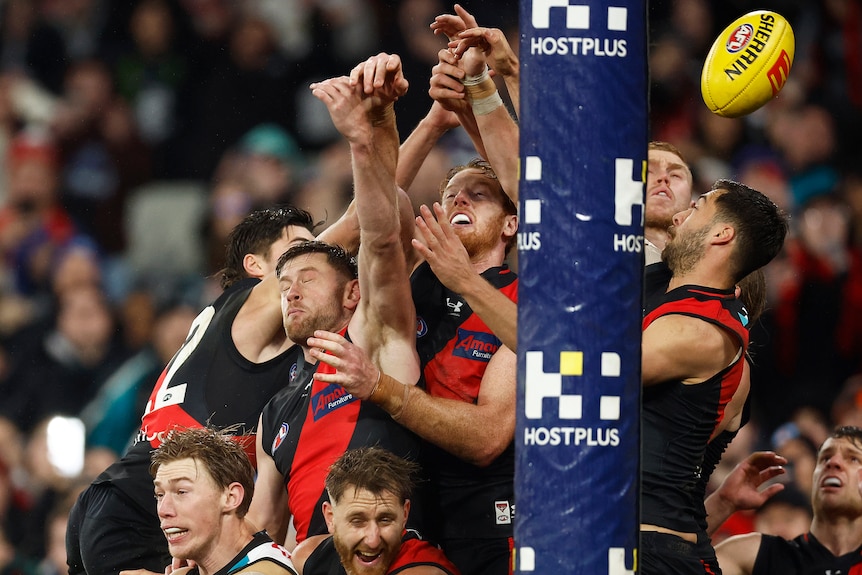 Essendon players grimace as they leap and try to punch the ball clear as it passes the goal line.