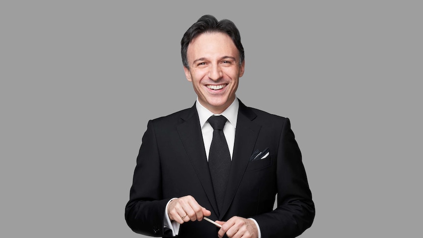 Conductor Umberto Clerici, wearing a black suit and holding a baton, stands in front of a grey background