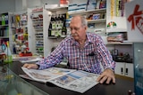 A man sitting behind a newsagent's counter reads a newspaper.