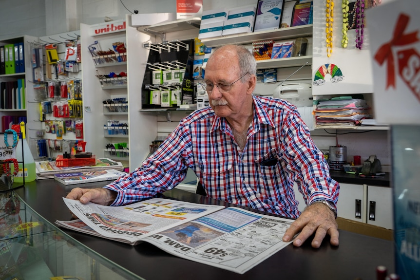 A man sitting behind a newsagent's counter reads a newspaper.