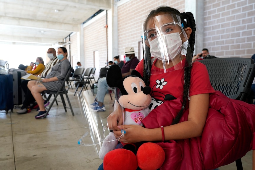 A girl with face mask and face shield on sits waiting