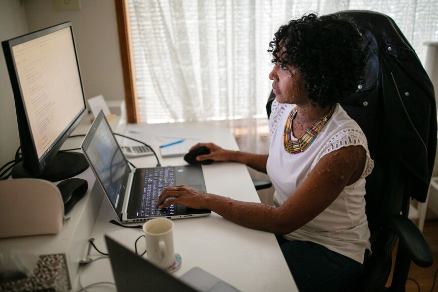 A woman in a white shirt sitting at a desk looking at a computer screen.