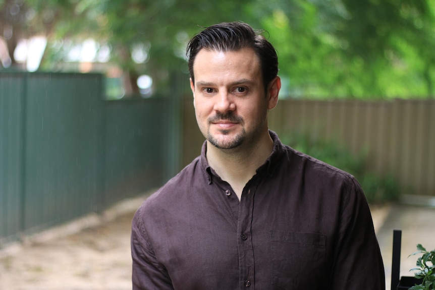 A man with short brown hair and a brown shirt standing outside by a fence