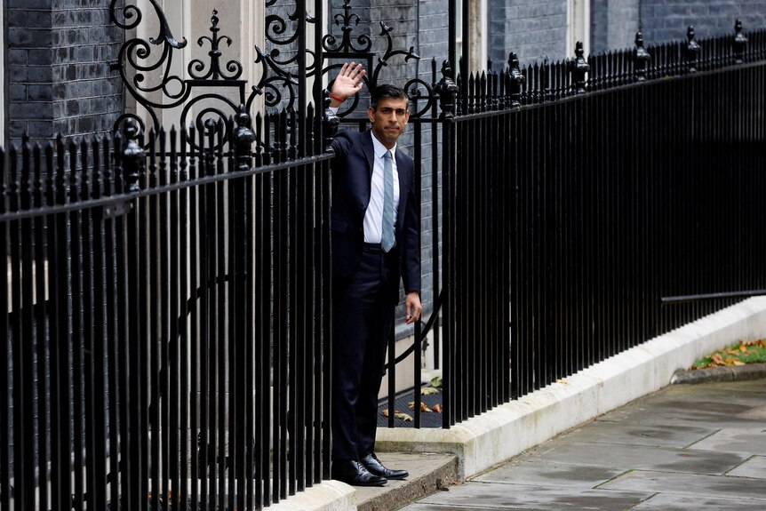 rishi sunak stands on the path at the gates of 10 downing street with his arm up waving