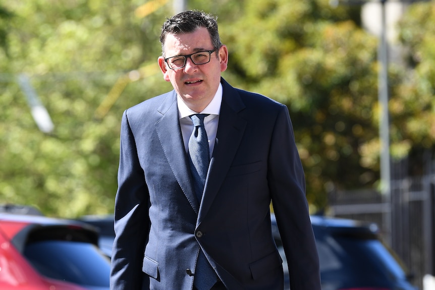 Daniel Andrews, dressed in a suit with briefcase and no mask, walks across the Parliament carpark on a sunny day.