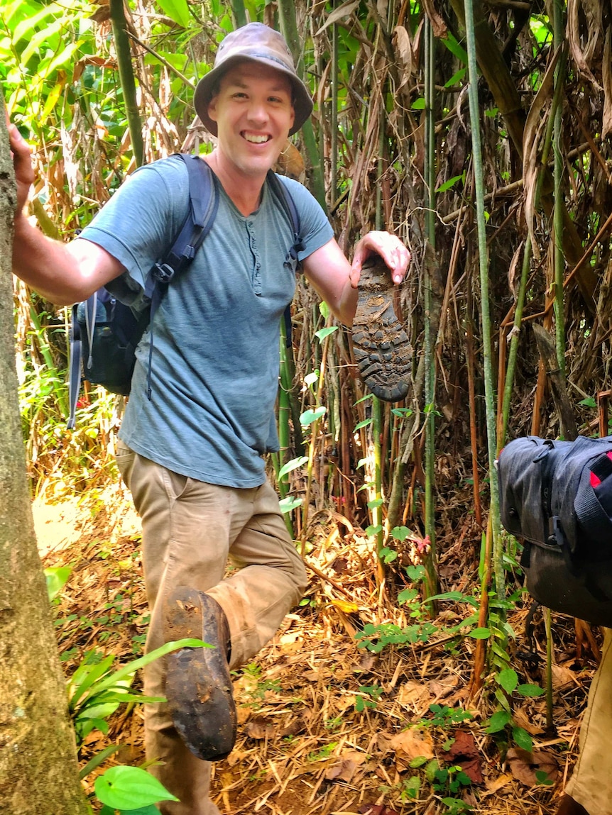 Harvey holding mud-caked sole of one boot that came off the rest of the boot while standing on one foot in jungle.