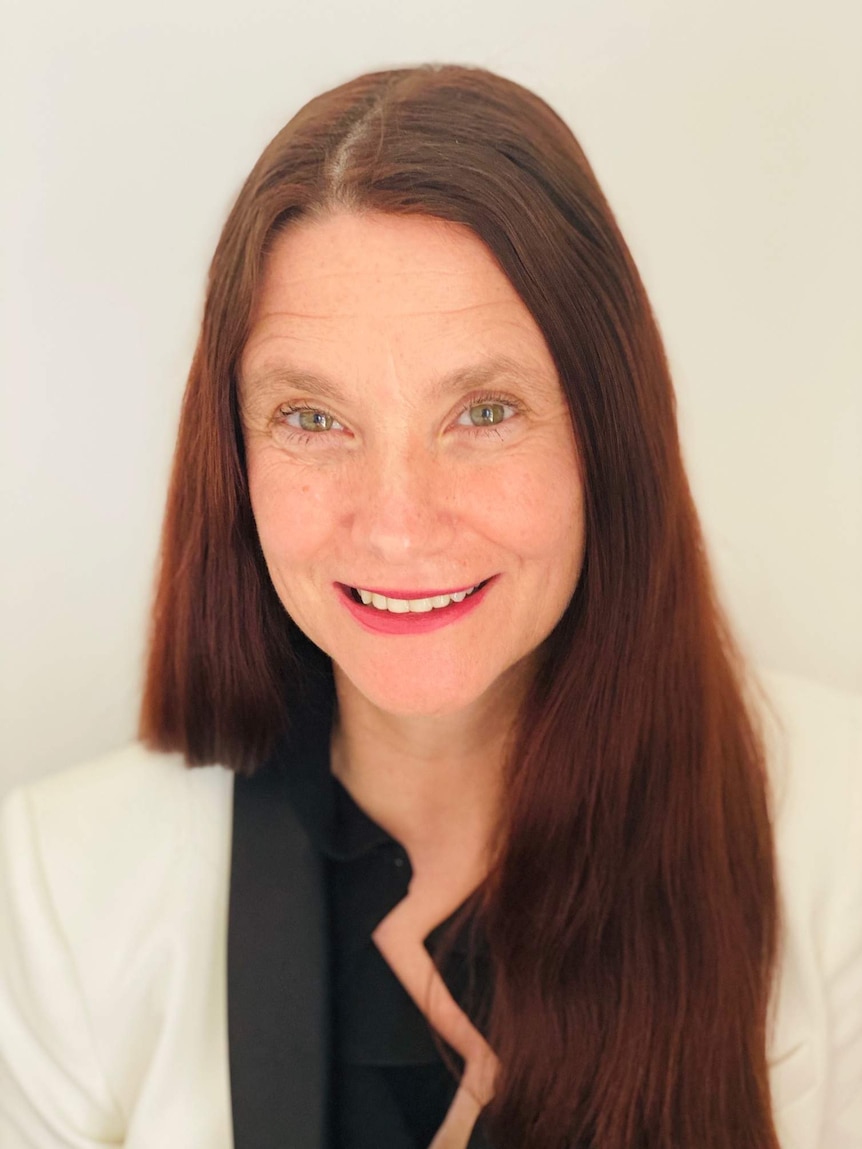 A headshot of a red haired woman wearing a black shirt and white blazer.