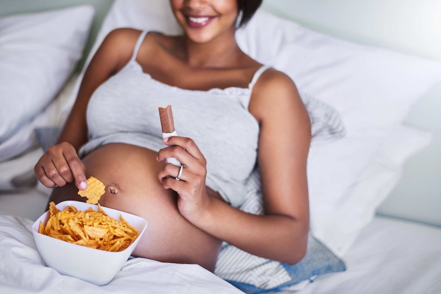 Pregnant woman lying in bed eating a chocolate bar and chips