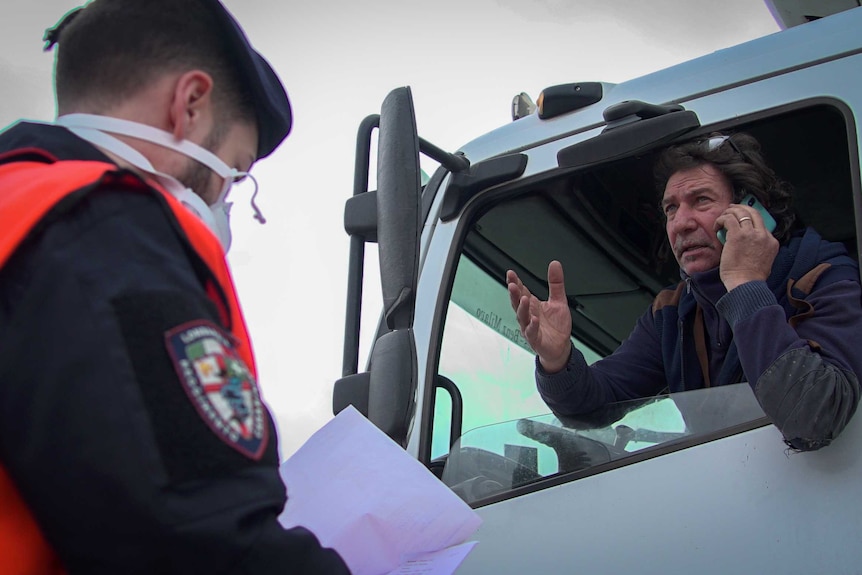 A leans out the window of his truck and gestures as a police officer checks his papers.