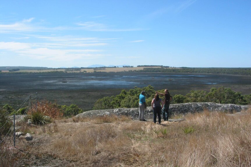 Wide shot of three people standing near a rocky outcrop overlooking wetlands and arid ranges