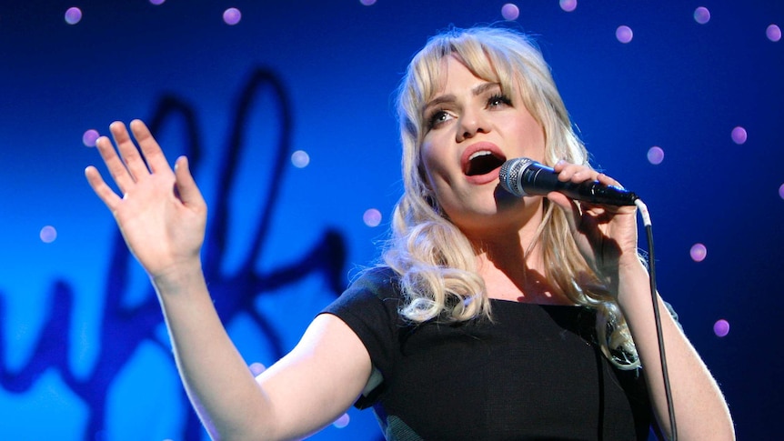 Blonde woman in a black top holds a microphone and sings on a stage.