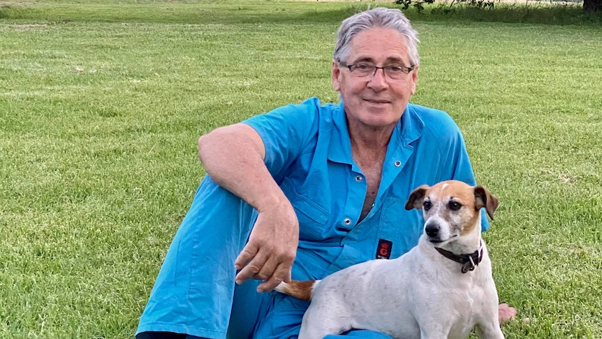 A man and his terrier sit on the grass and smile at camera.
