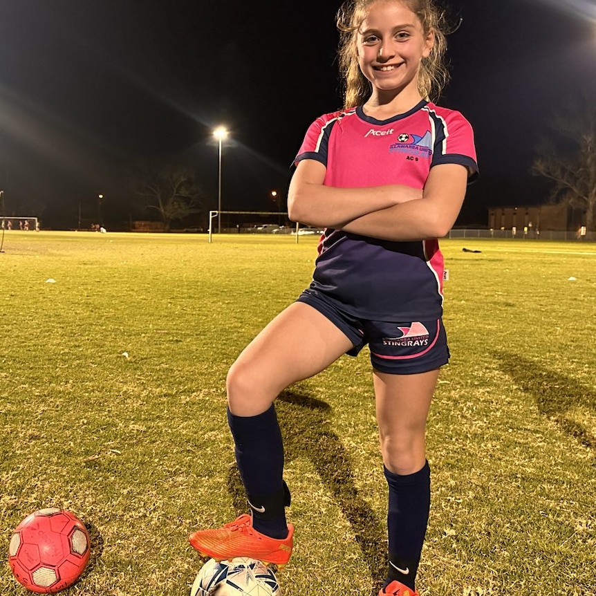 Young girls in a soccer uniform stands with arms folded and foot on the ball 