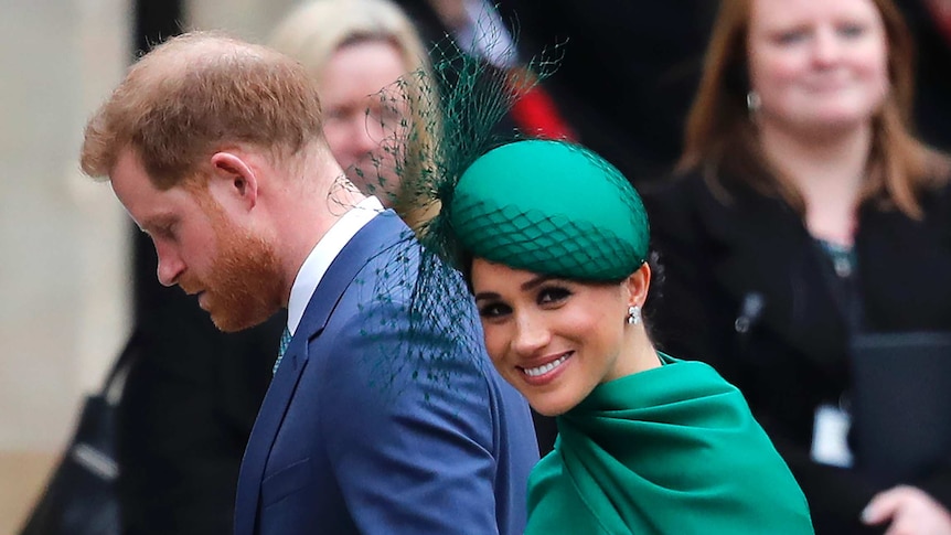 Britain's Prince Harry, wearing a suit and tie, and Meghan in a green dress walk together as Meghan looks toward the camera.