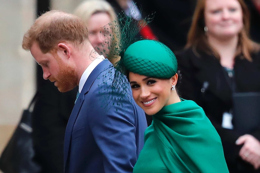 Britain's Prince Harry, wearing a suit and tie, and Meghan in a green dress walk together as Meghan looks toward the camera.