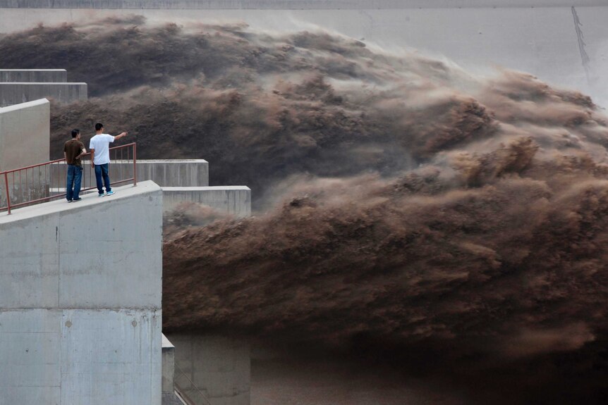 Two men stand atop large concrete dam pylons as an enormous body of brown water gushes past them.
