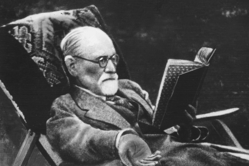 Sigmund Freud, wearing glasses and with a short grey beard, reads a book in a deck chair in the garden.
