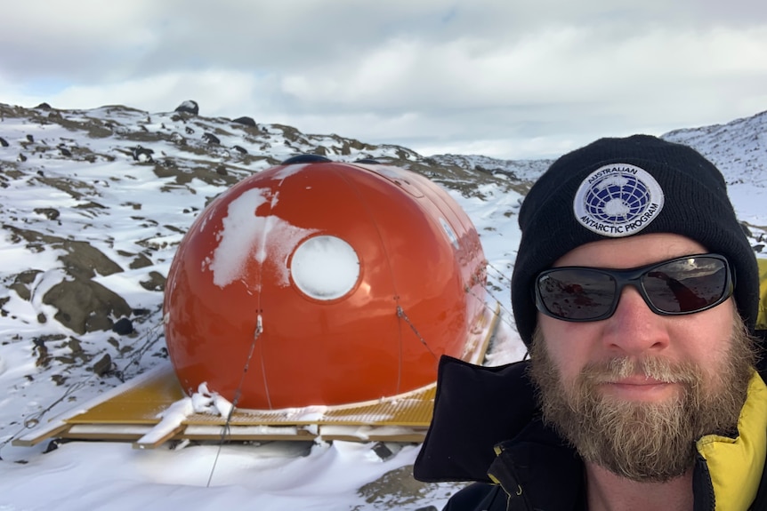 David wearing thick cold weather clothes and a beanie, on icy ground in Antarctica with a red object behind.