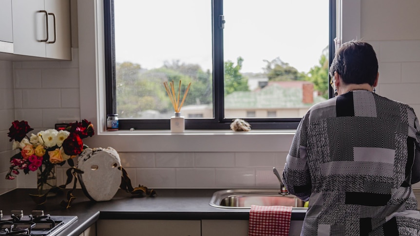 A woman with short brown hair wearing a grey patterned shirt does dishes at a sink below a window with her back the camera.