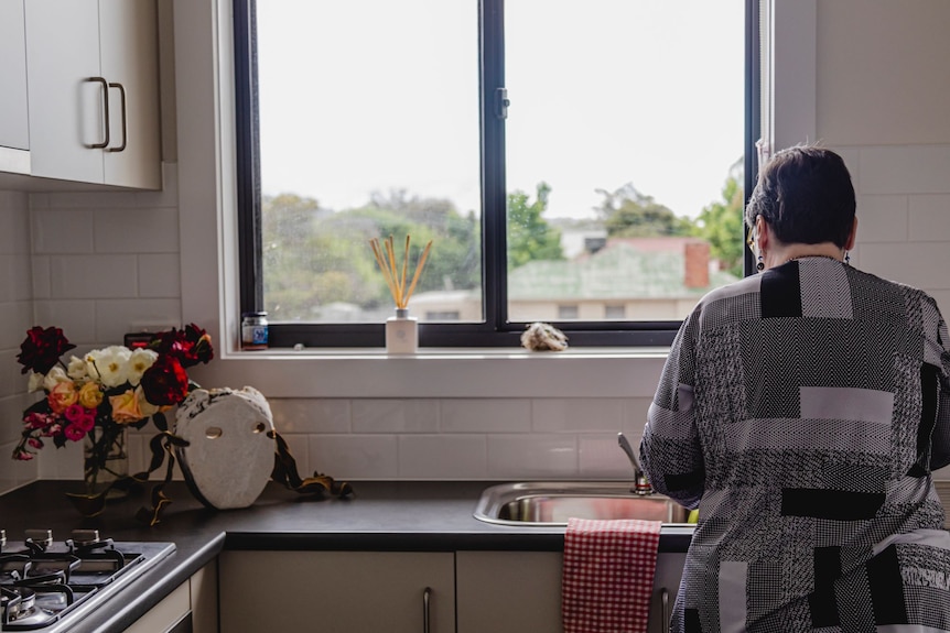 A woman with short brown hair wearing a grey patterned shirt does dishes at a sink below a window with her back the camera.