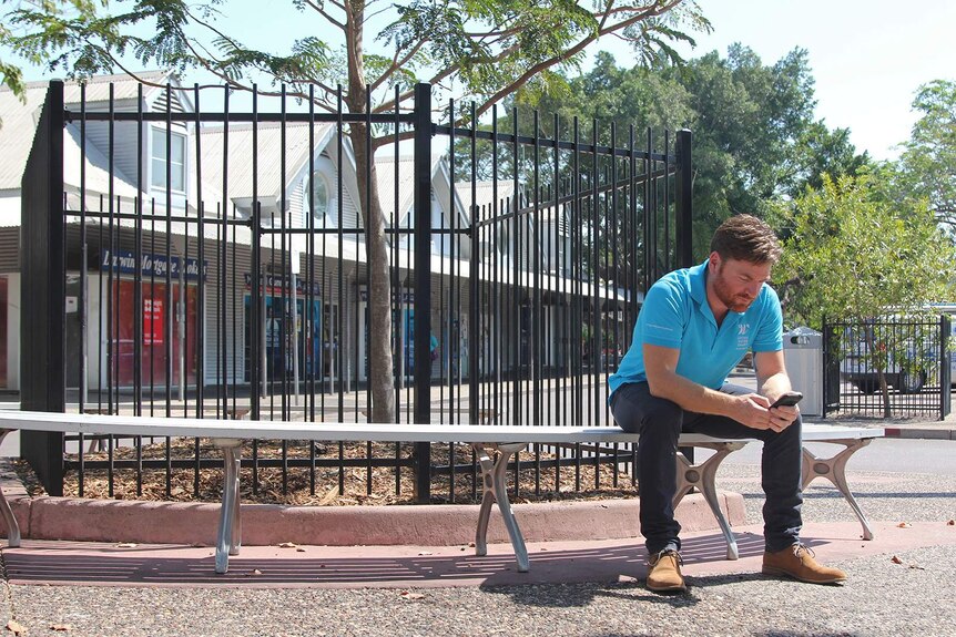 A photo of Shane Burgess sitting on a bench in Parap texting on his phone.