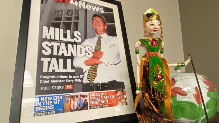 A framed newspaper front page featuring a smiling Terry Mills, sitting on a book shelf, next to a souvenir Indonesian figure