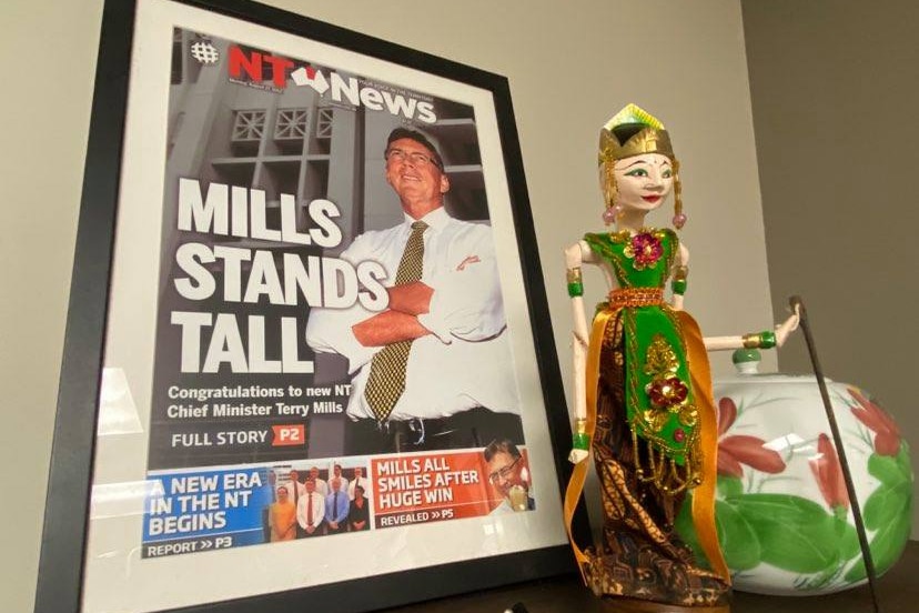 A framed newspaper front page featuring a smiling Terry Mills, sitting on a book shelf, next to a souvenir Indonesian figure