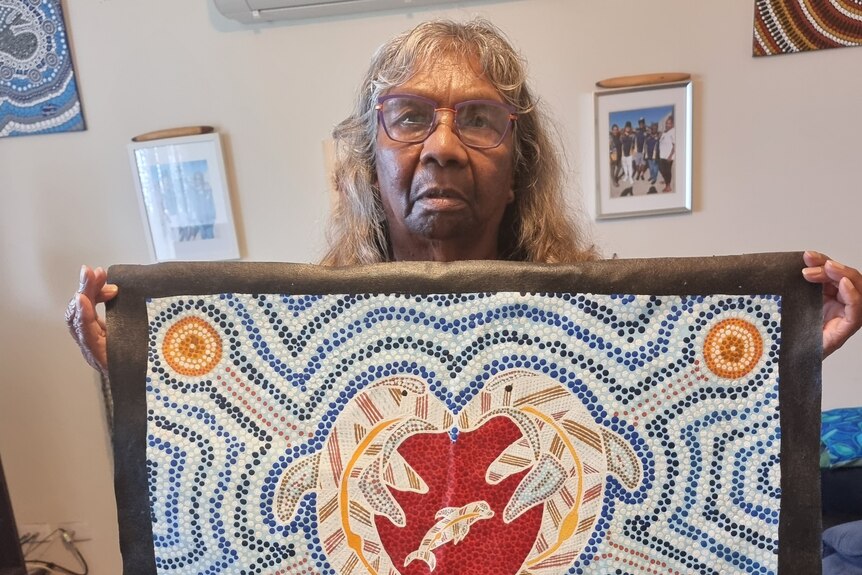 A woman stands in a lounge room holding up an indigenous dot painting. 