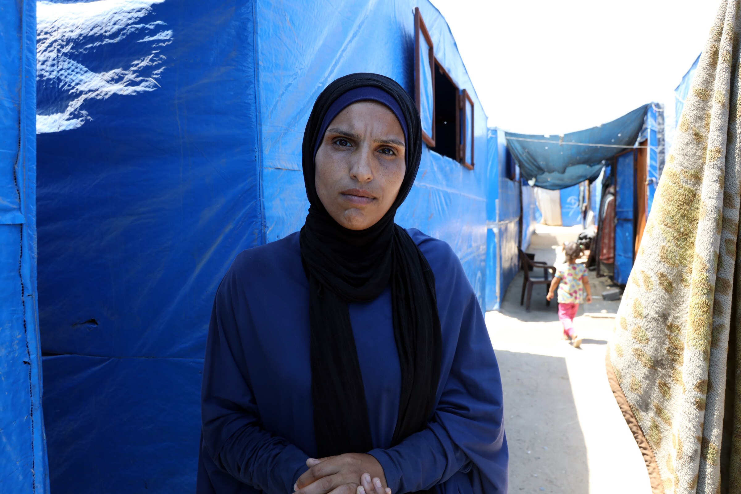 Umm Fadi clasps her hands as she stands in shade between rows of tarpaulin tents.