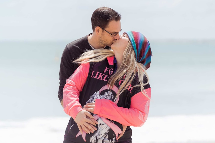 A mid shot of a man and a woman facing the camera as they hug and kis on a beach.