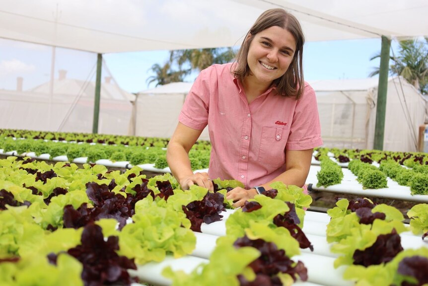 A young white woman, Zoe, with shoulder length brown hair smiles wearing a red chequered work shirt as she tends to lettuce.