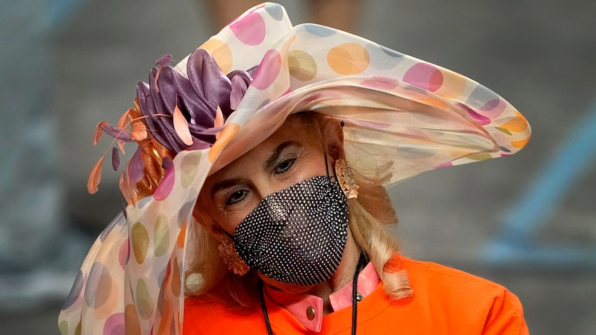 A woman in a big hat, orange shirt and covid mask looks at a clip board 