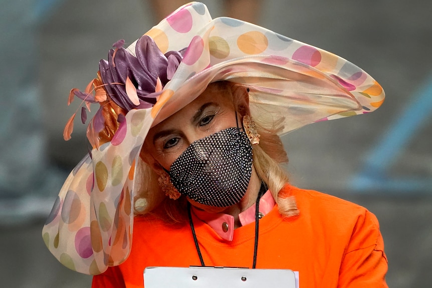 A woman in a big hat, orange shirt and covid mask looks at a clip board 