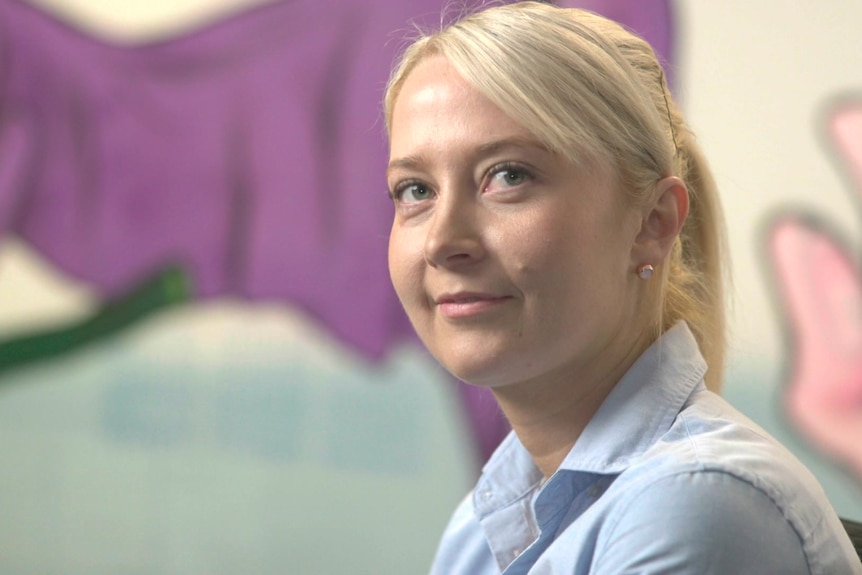 A blonde woman sits smiling, a colourful painted wall behind her.