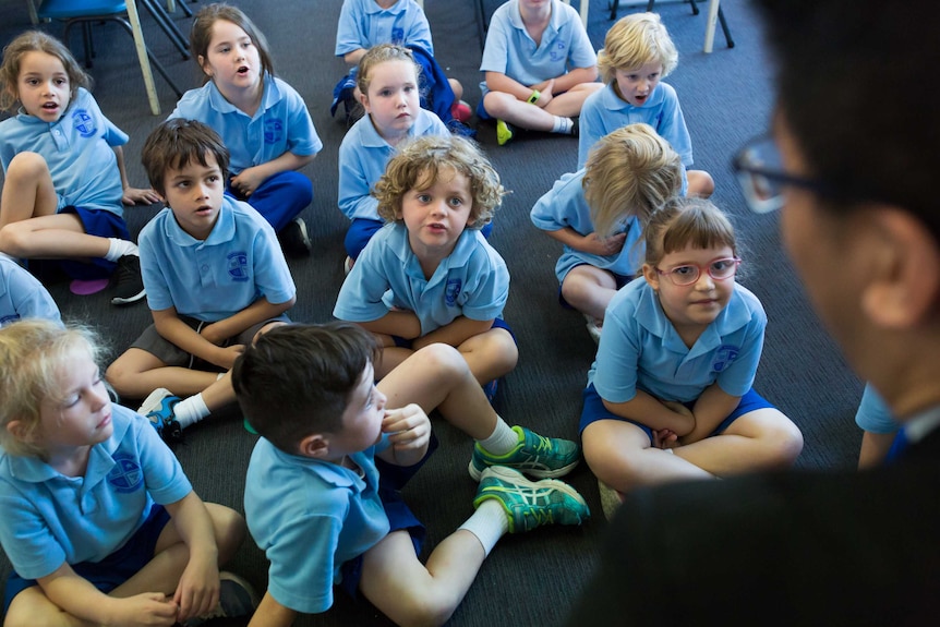Children in various states of squirm and fidget sit cross-legged on the floor, eyes focussed on Father Justel Callos.