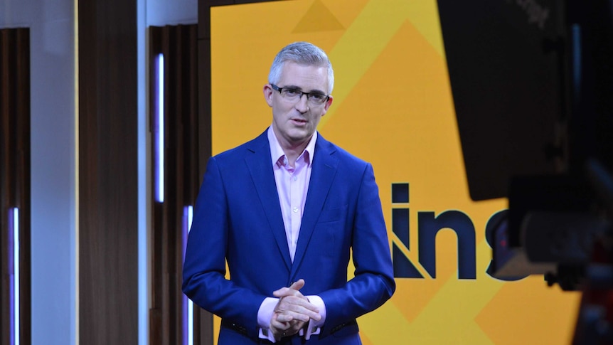 Speers standing in front of yellow TV screen in studio looking to camera.