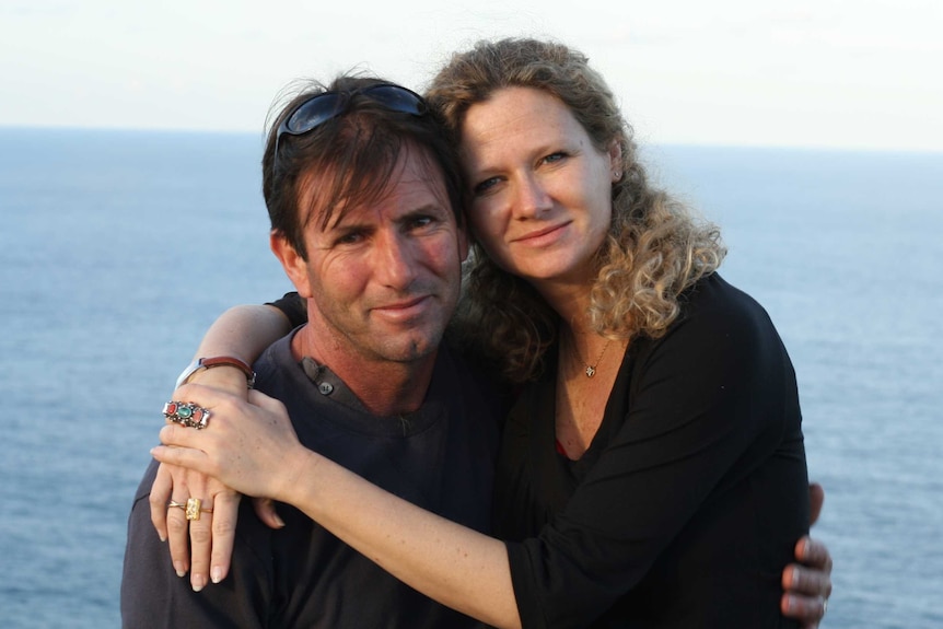 Benedetta and her husband Brian stand in front of the ocean. They have their arms around each other and are smiling.