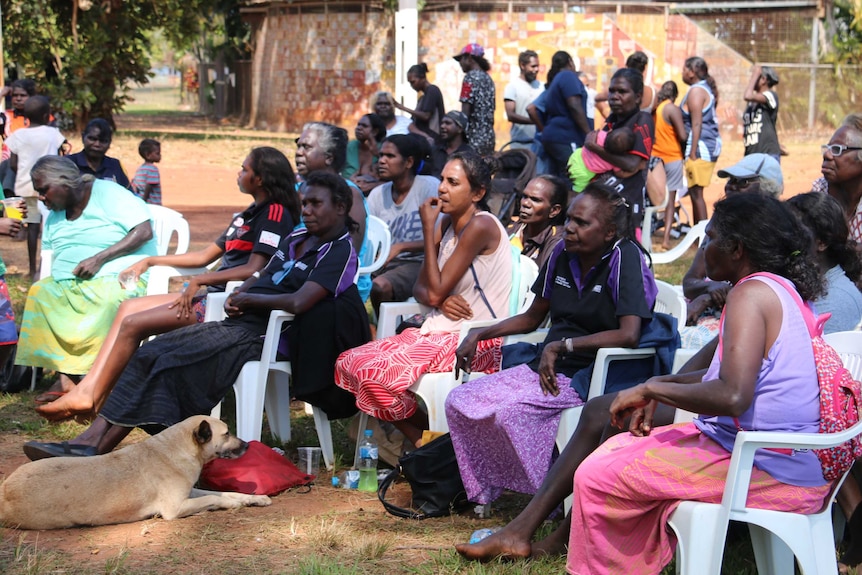 A crowd of Indigenous women sit in chairs outside, looking on.