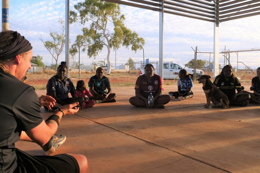 A white man sits as part of a circle with Aboriginal women under a shade structure. They are all smiling and engaged.