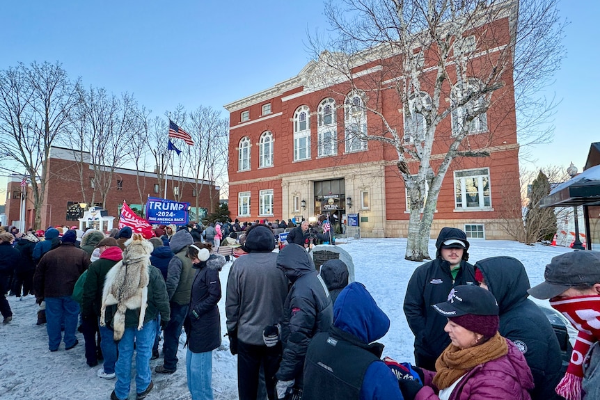 People queue in the snow outside a red brick building. 