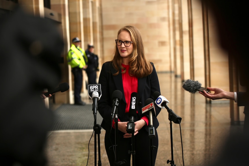 A woman wearing a red shirt, black blazer, and glasses stands before mircrophones outside of WA Parliament