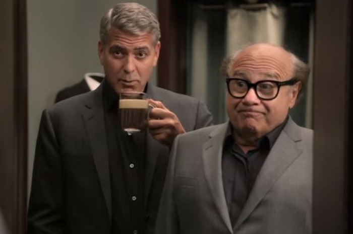 A man wearing a suit and drinking a cup of coffee looks in a mirror behind a shorter, older man.
