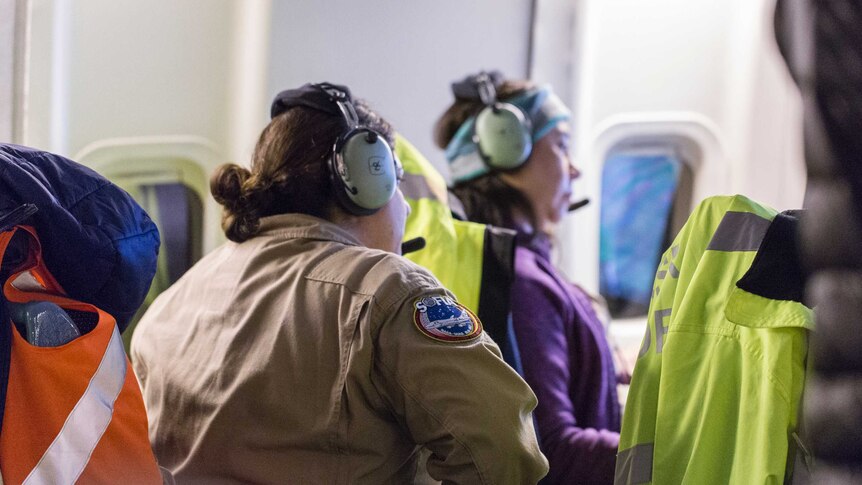 Women in an aircraft cabin wearing headsets and NASA-badged jumpsuits and vests stare intently in the same direction.