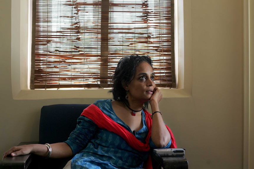 Roy sits in a a chair, in front of a blinded window, wearing blue and pink.